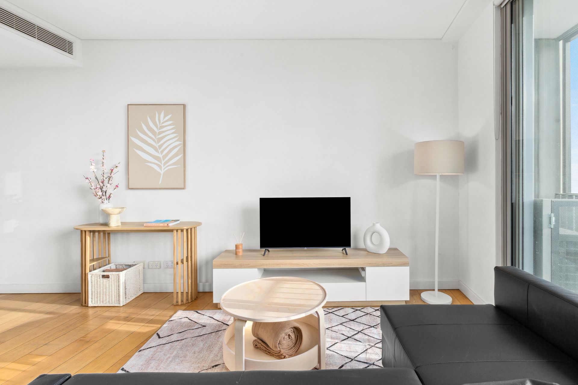 Minimalist living room with white decor, black sofa, TV, wooden console, and floor lamp by a window.