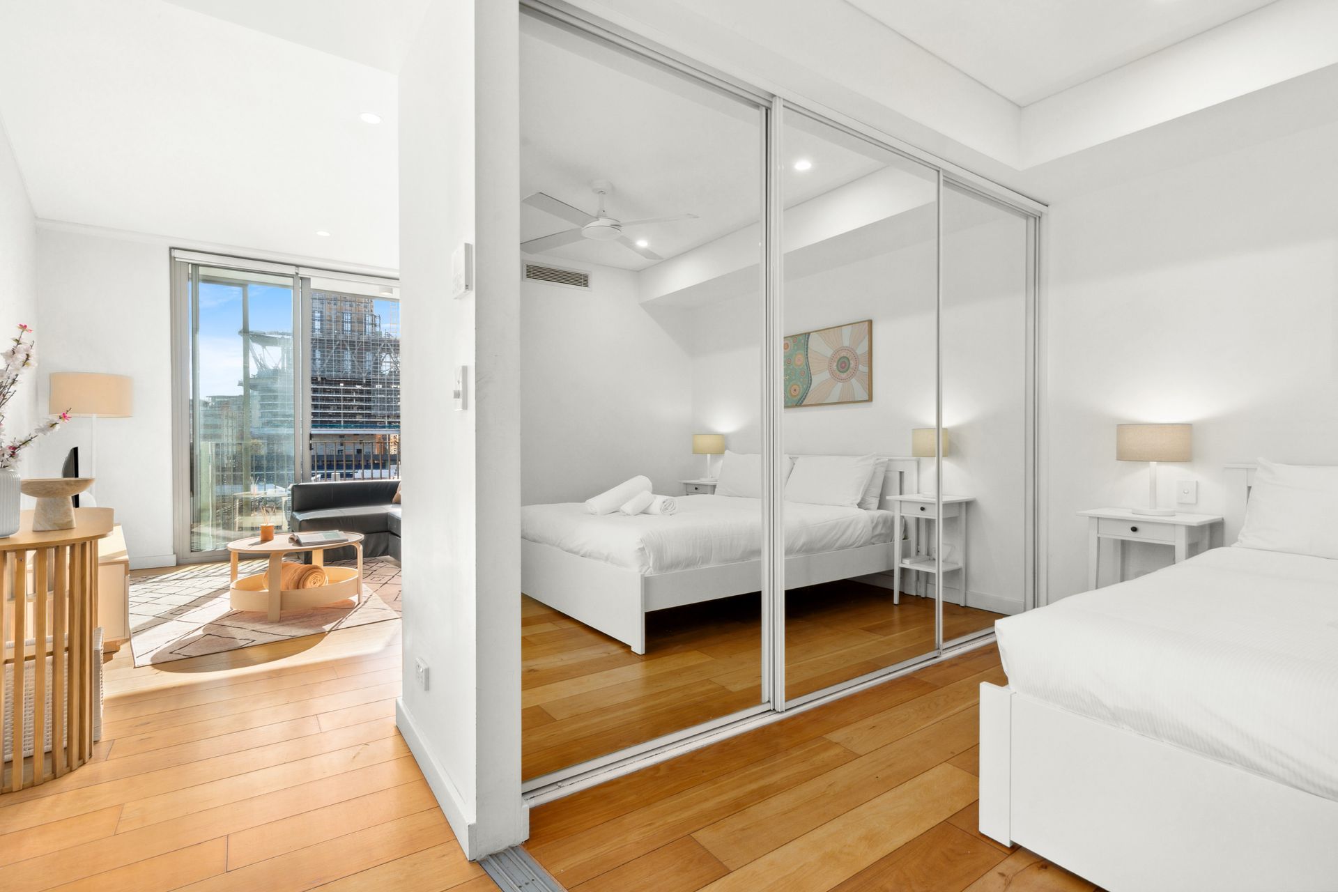 Bright bedroom with mirrored closet doors, white furniture, and a sunlit city view through open balcony doors