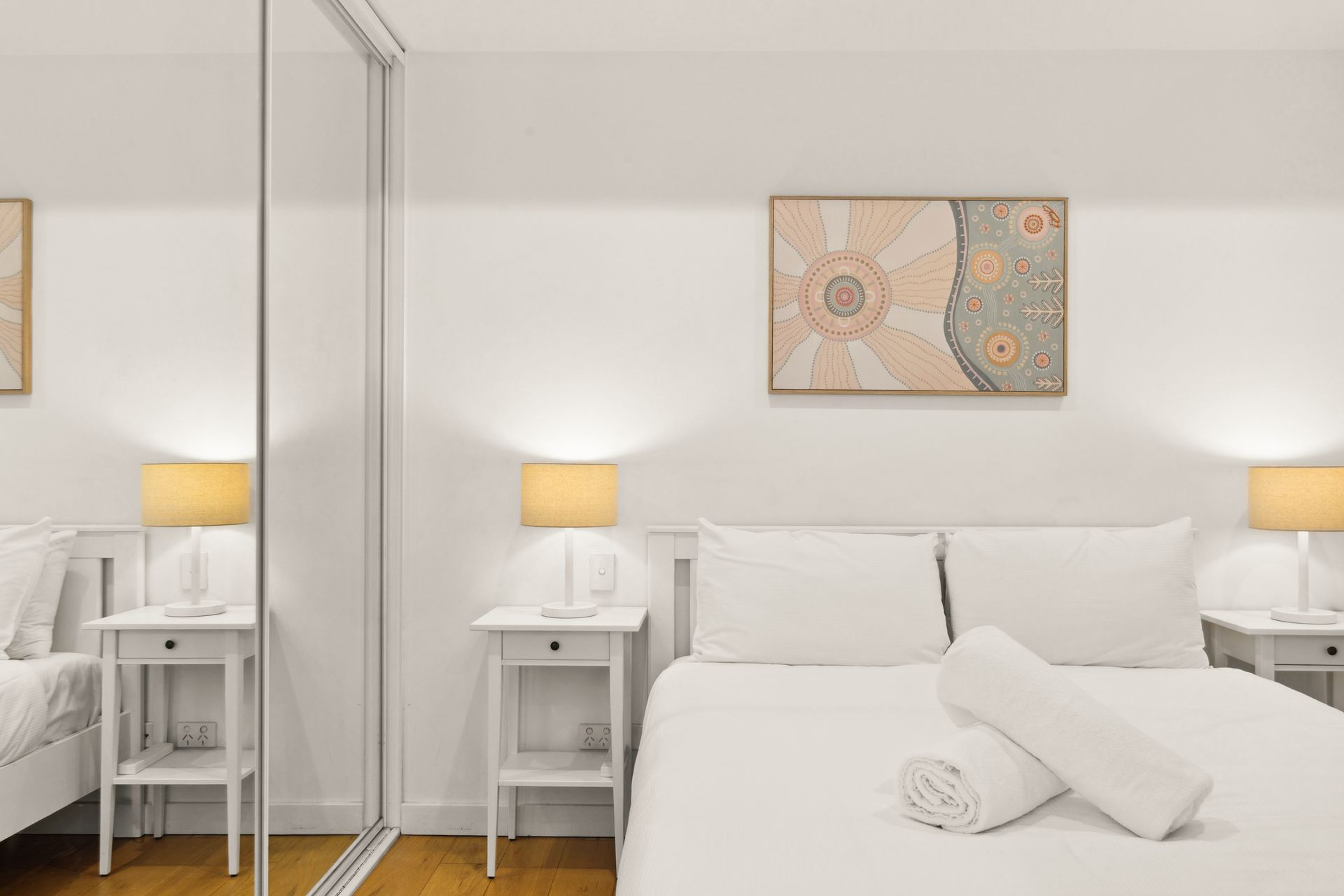 Bright white bedroom with a bed, bedside lamps, mirrored wardrobe, and abstract wall art