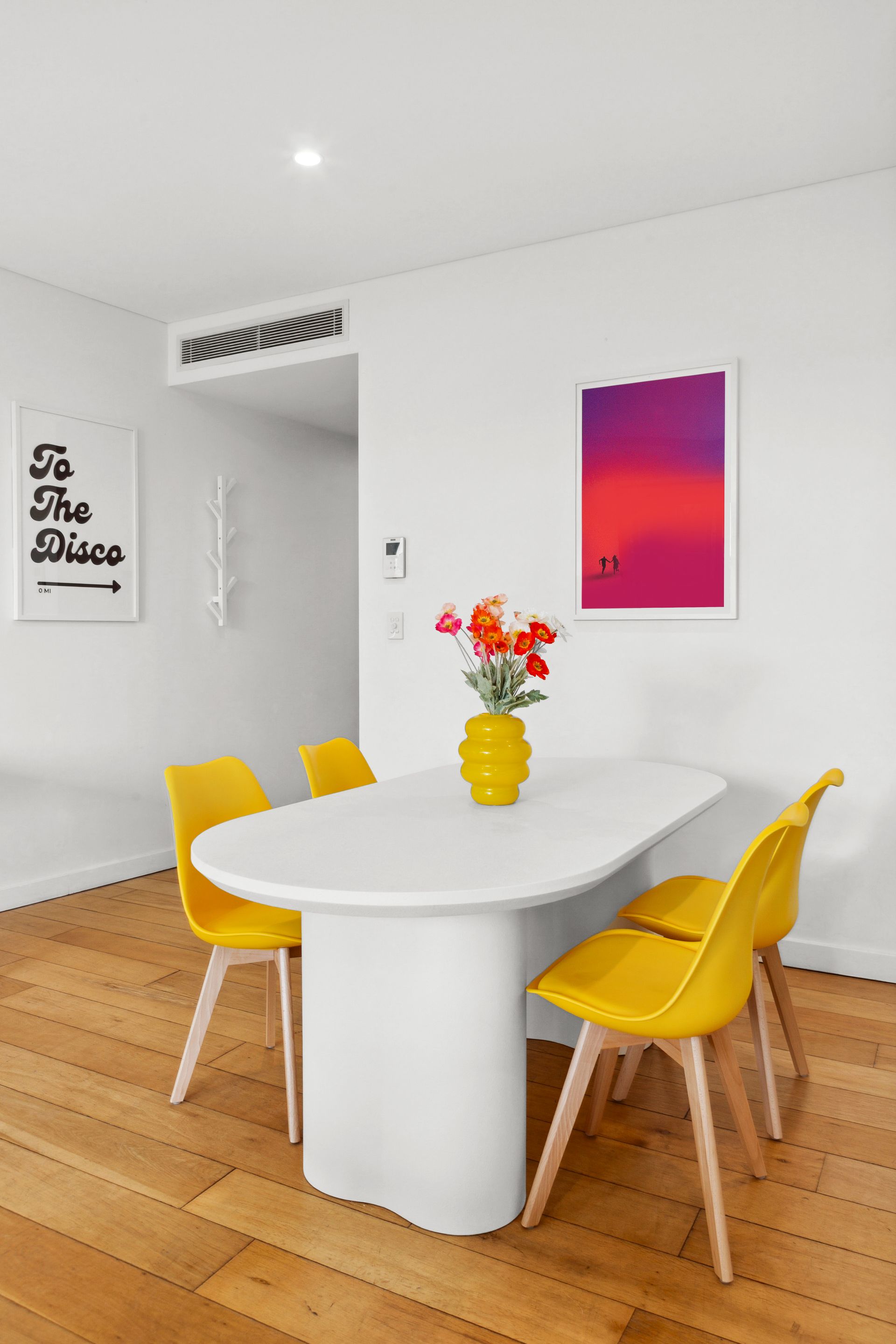 Bright dining room with white oval table, yellow chairs, wood floor, and colorful wall art and flowers.