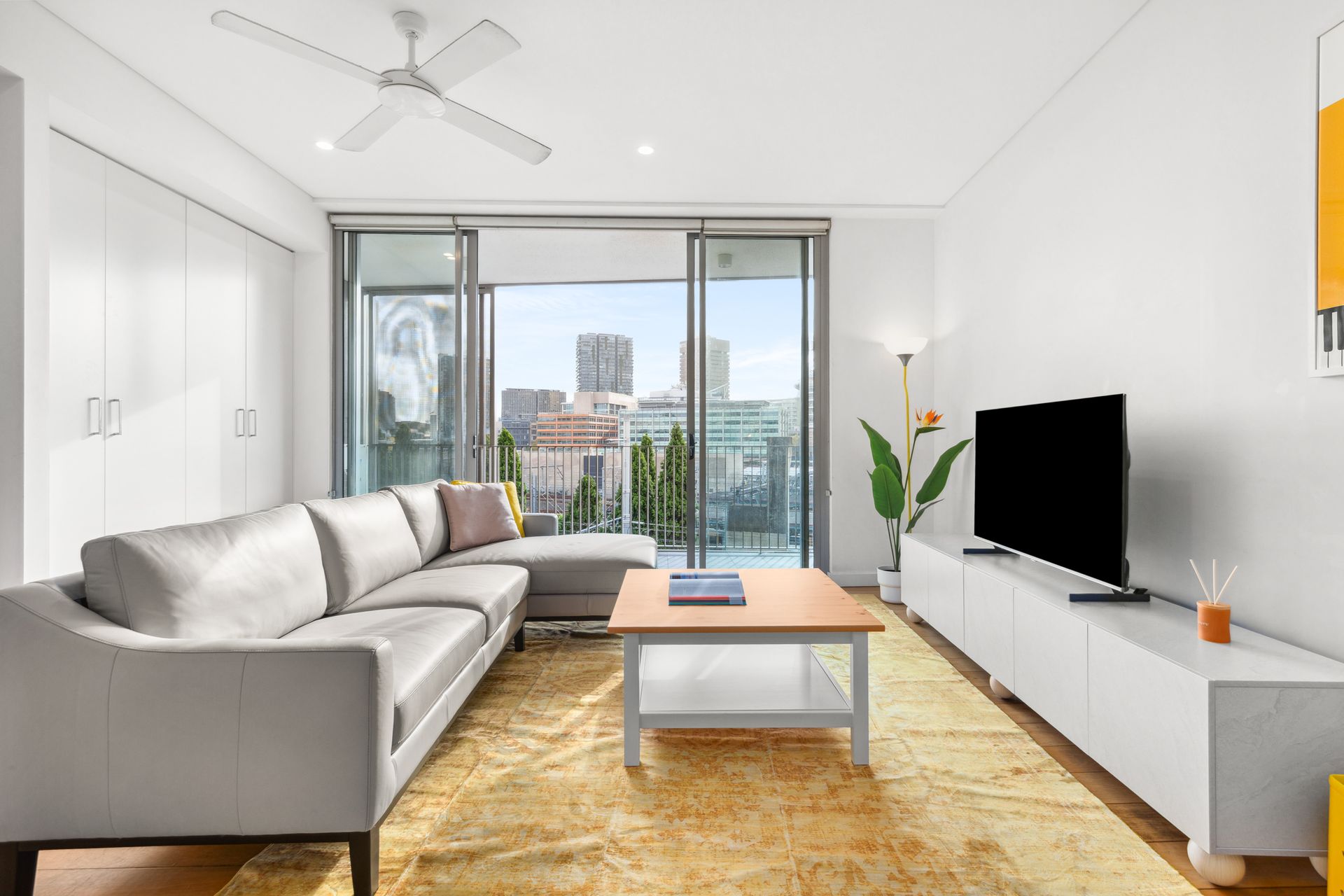 Bright living room with gray sectional, TV, yellow rug, and city-view balcony doors