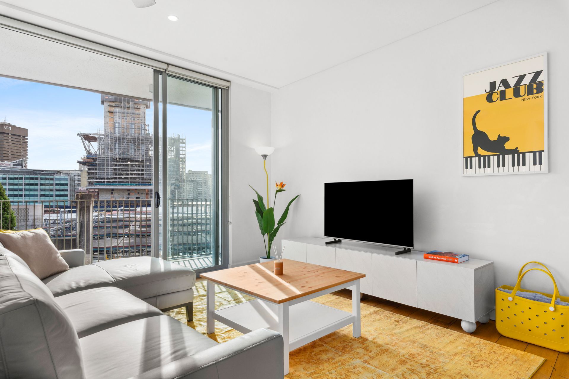 Bright modern living room with sofa, TV, glass coffee table, city view, and yellow accent chair