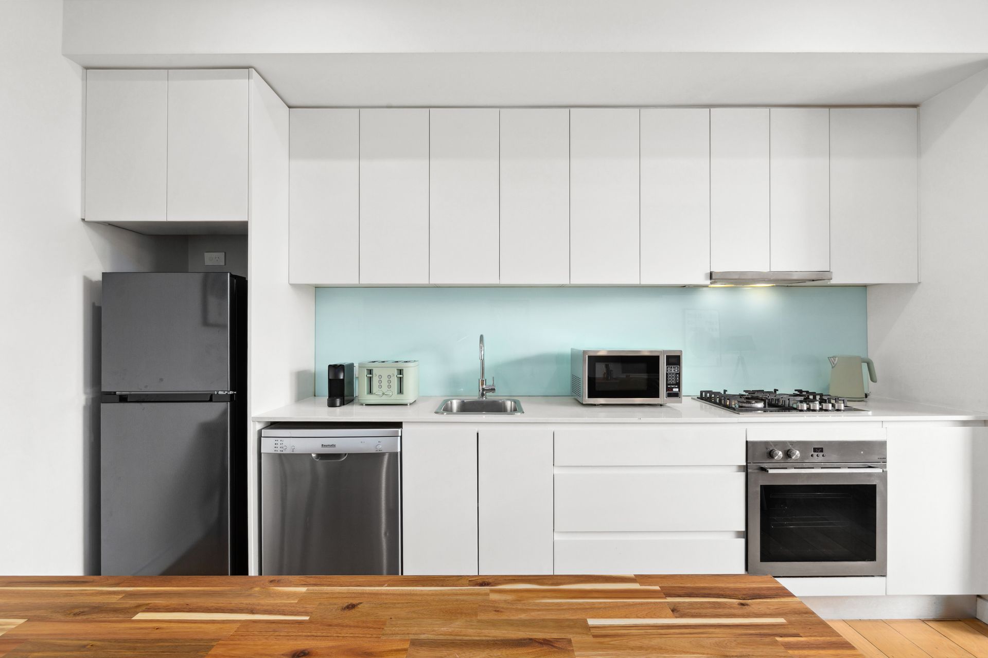 Modern white kitchen with stainless appliances, pale blue backsplash, and wooden table in foreground