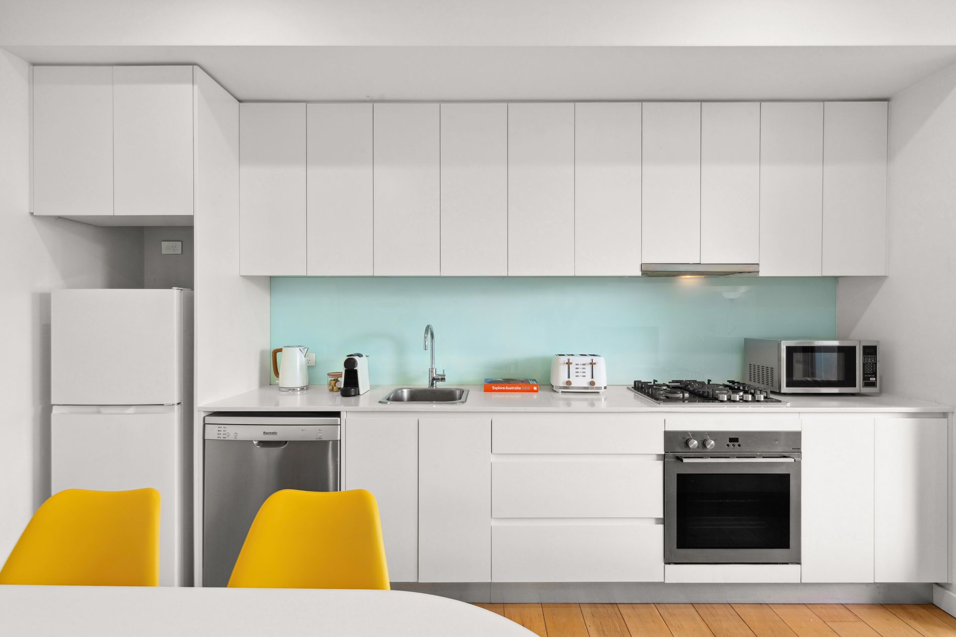 Modern white kitchen with yellow chairs, pale blue backsplash, and stainless steel appliances