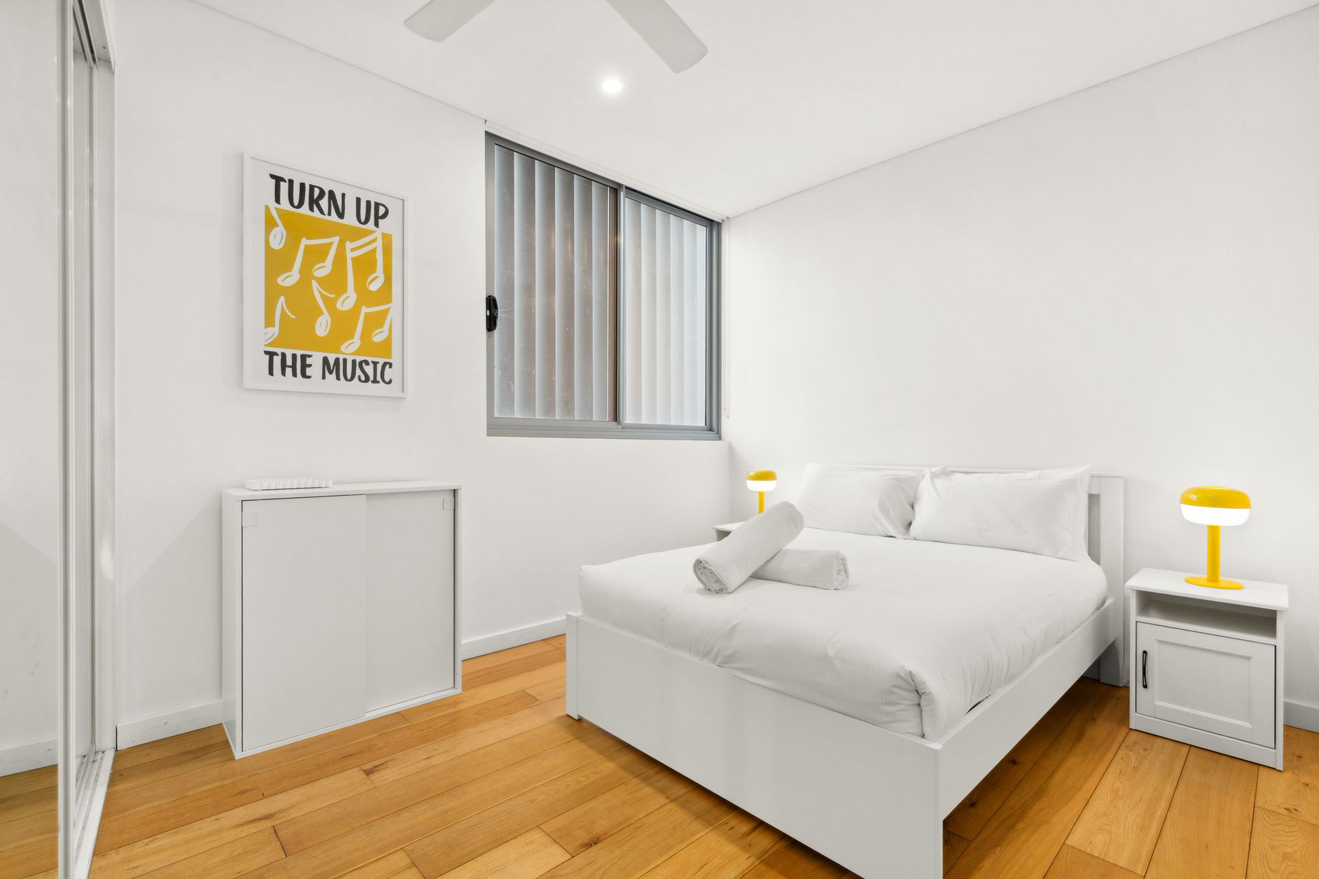 Bright white bedroom with wooden floor, twin bed, yellow lamp, dresser, and wall poster.