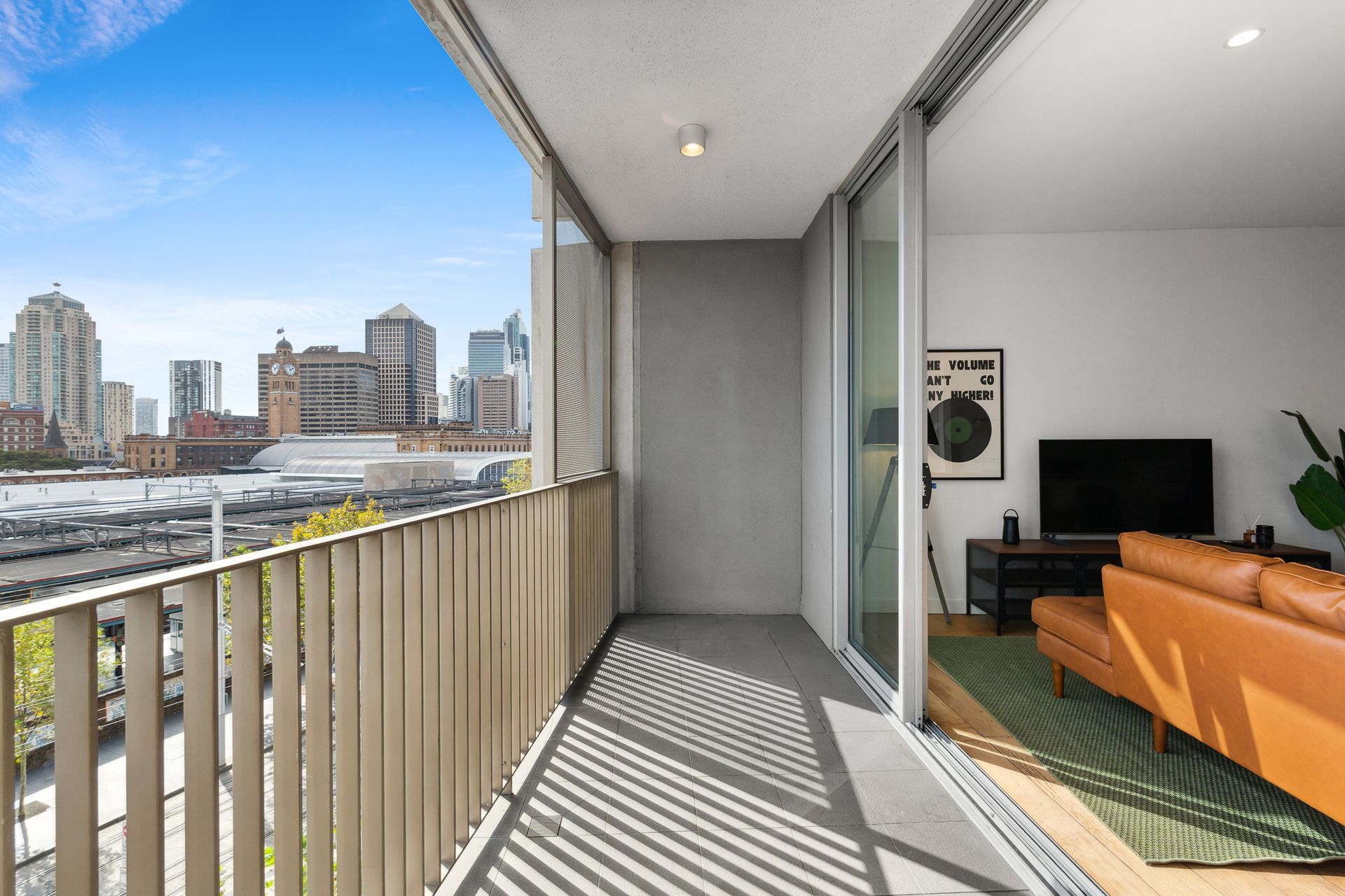Balcony overlooking city skyline, with orange sofa and TV visible in a modern apartment living room.