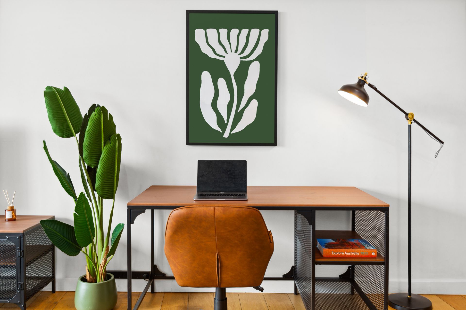 Minimal home office with desk, laptop, orange chair, tall plant, floor lamp, and wall art