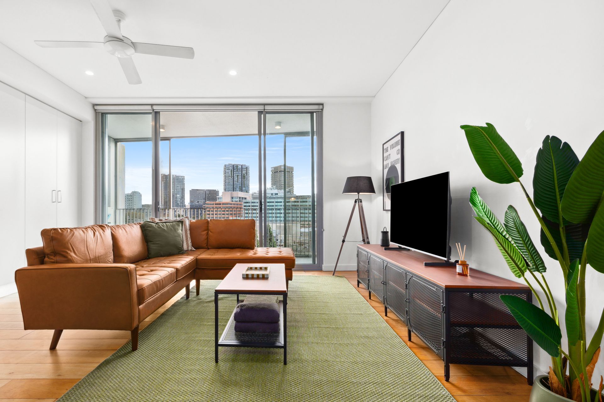 Modern living room with brown sectional, green rug, TV, large plant, and balcony city view