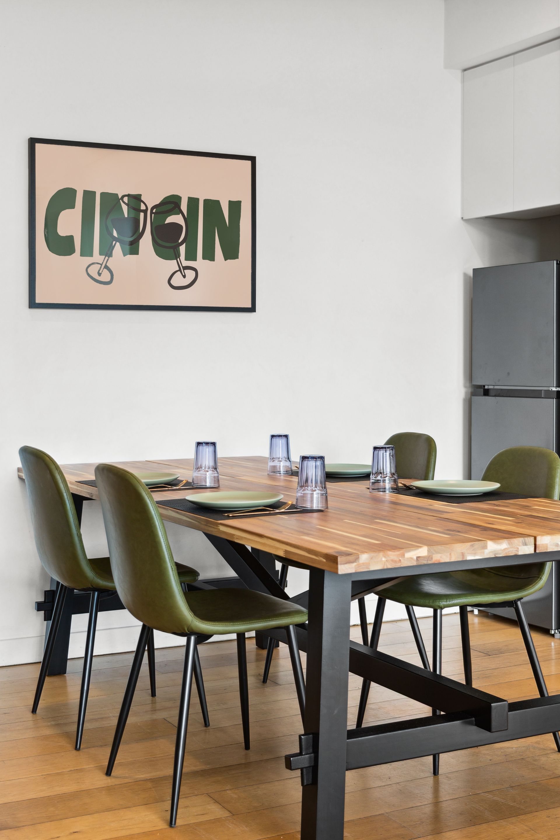 Dining room with a wooden table, green chairs, place settings, and a framed wall print near a kitchen.