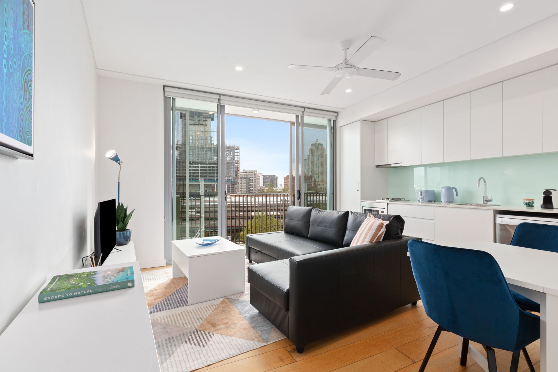 Bright modern living room with black sofa, blue chair, white kitchen, and balcony city view