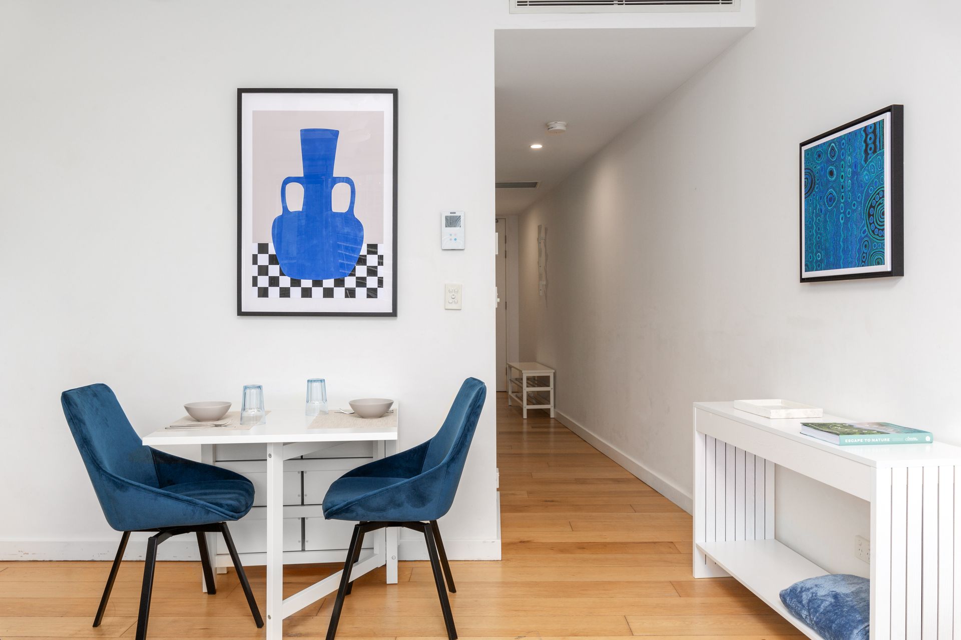 Bright dining nook with blue chairs, white table, and framed art in a minimalist hallway seating area