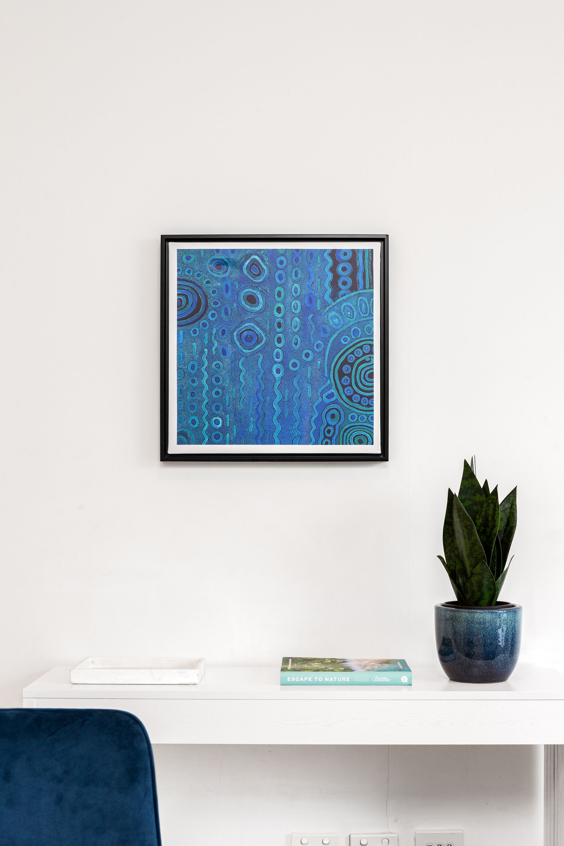 Minimal white shelf with blue abstract wall art, books, and a potted plant; teal chair edge at left.