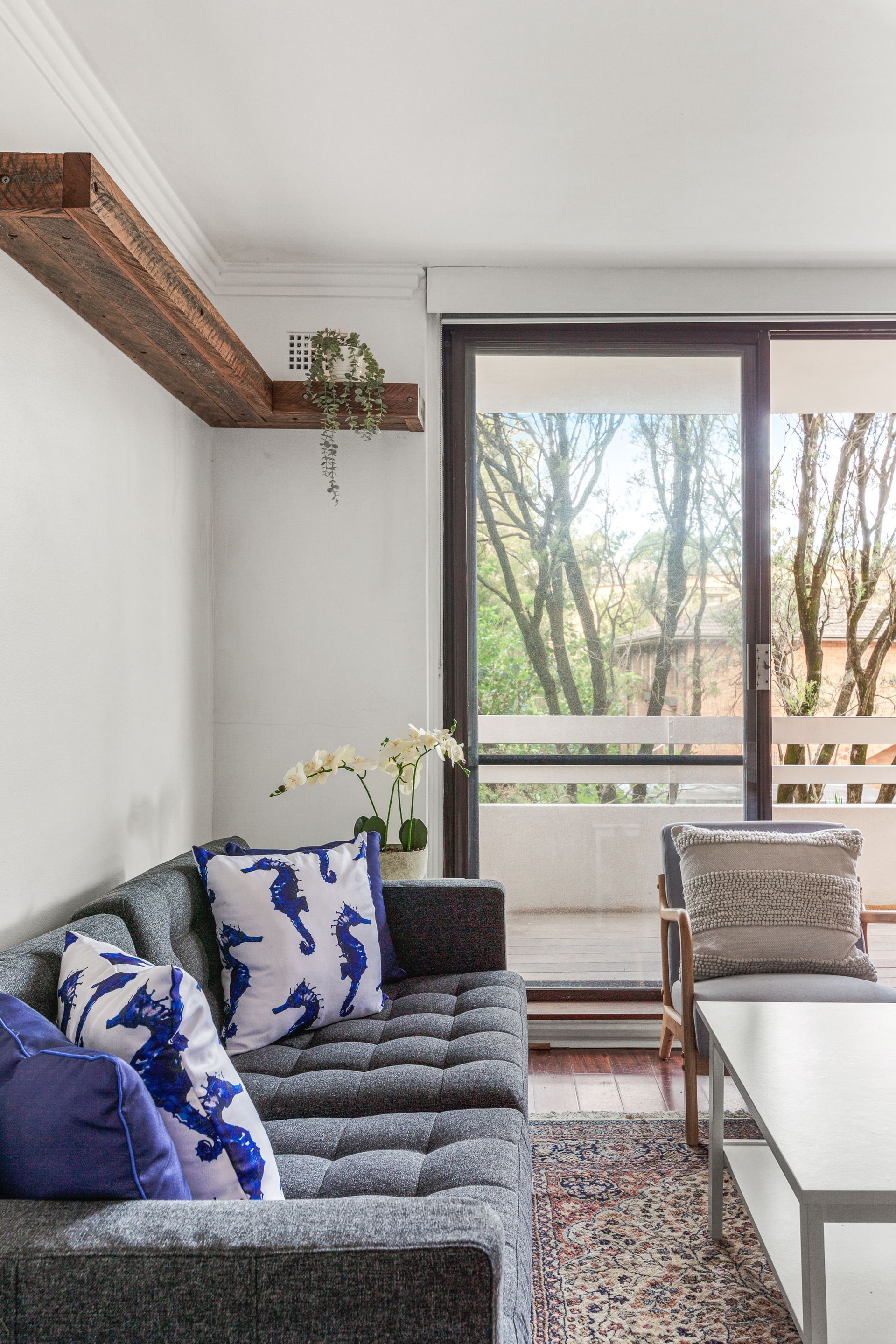 Modern living room with gray sofa, blue pillows, coffee table, and large window with trees outside