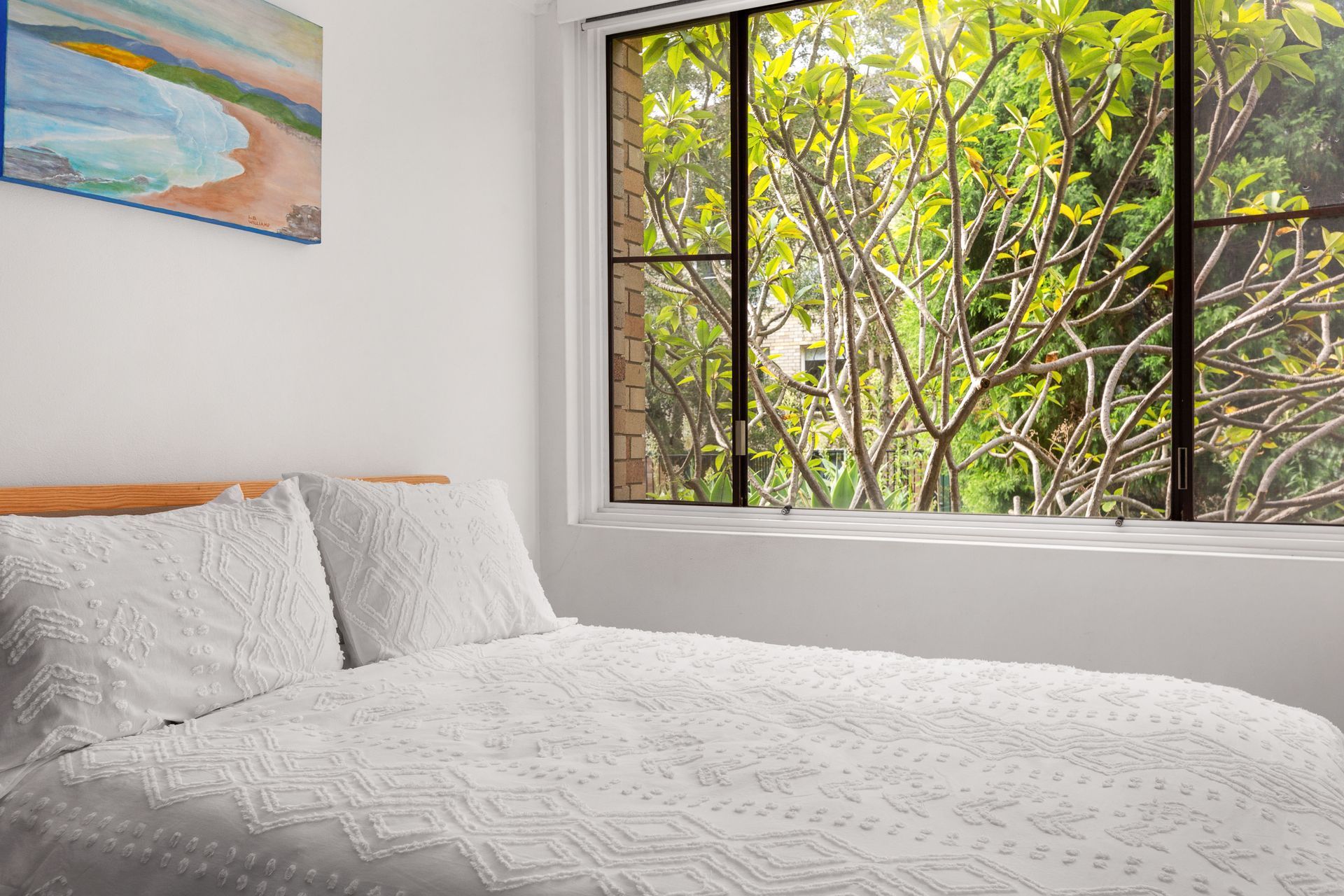 Bright bedroom with white bed and pillows beside a large window overlooking leafy green trees