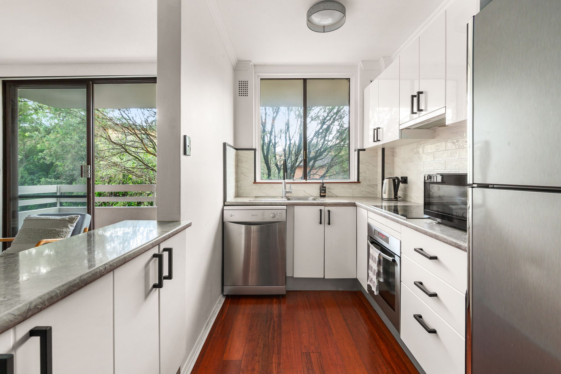 Bright modern kitchen with white cabinets, stainless appliances, and a large window overlooking greenery
