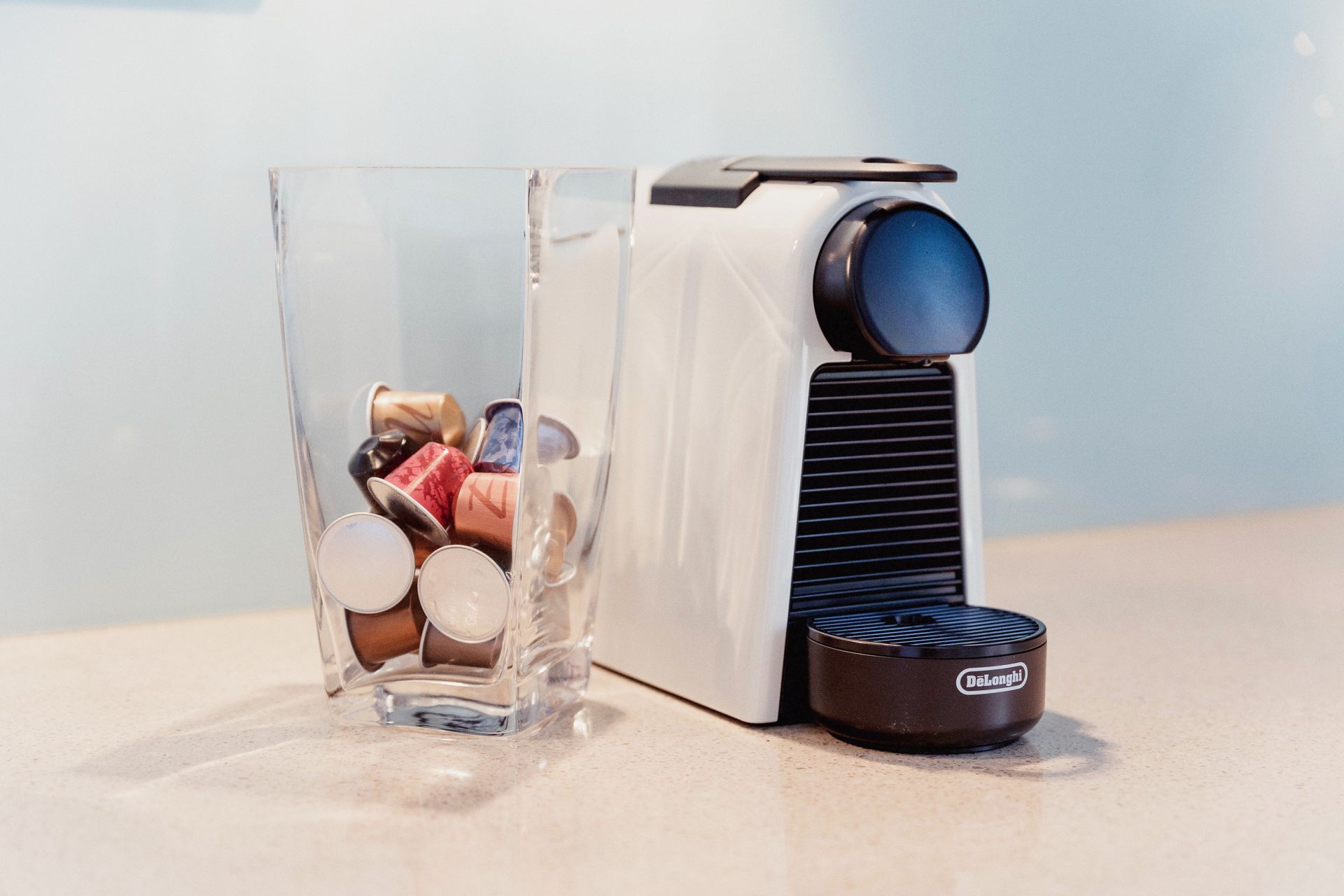 Single-serve coffee machine beside a clear cup of assorted coffee pods on a white surface