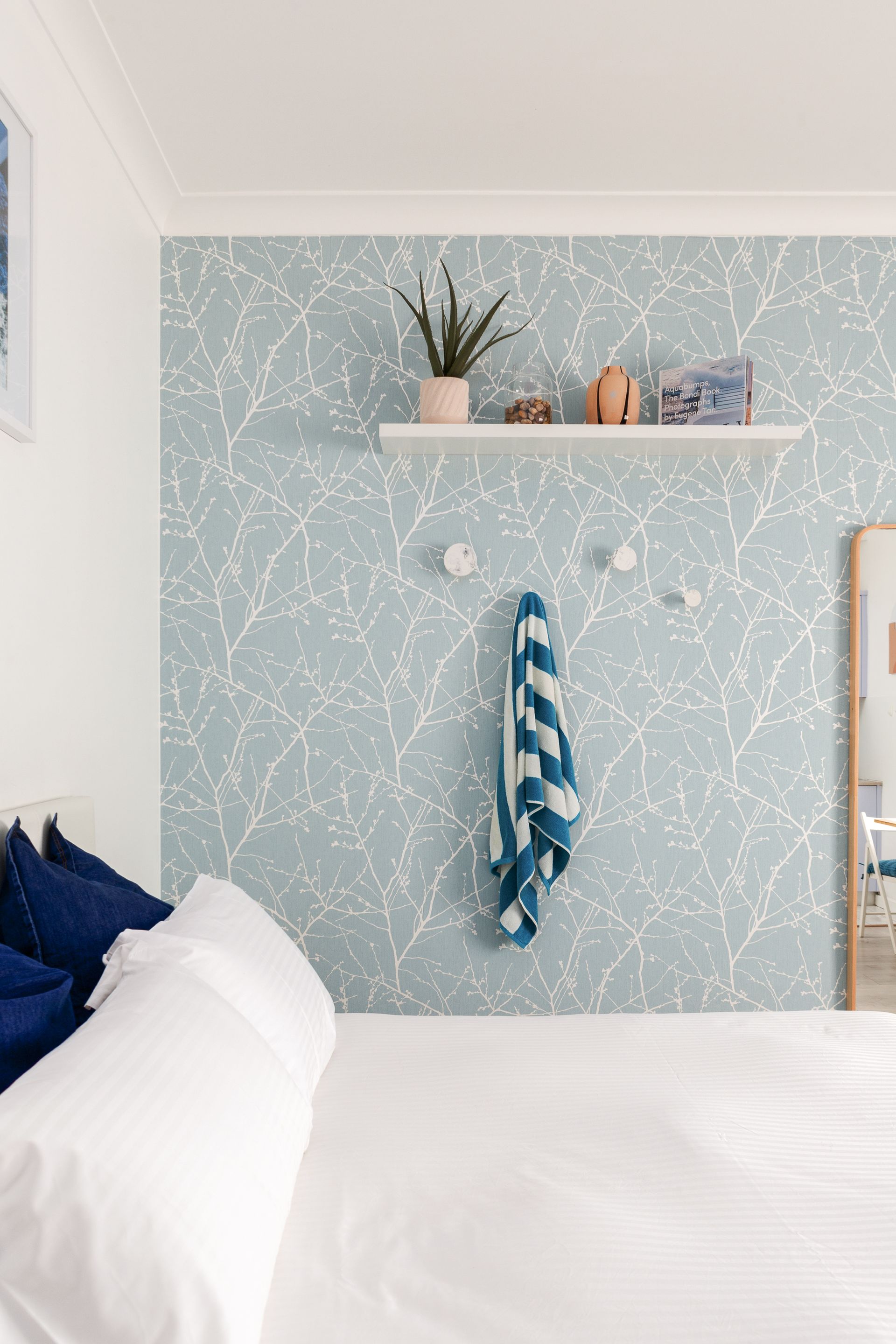 Cozy bedroom with blue patterned wall, white bed, and shelf with plants and decor