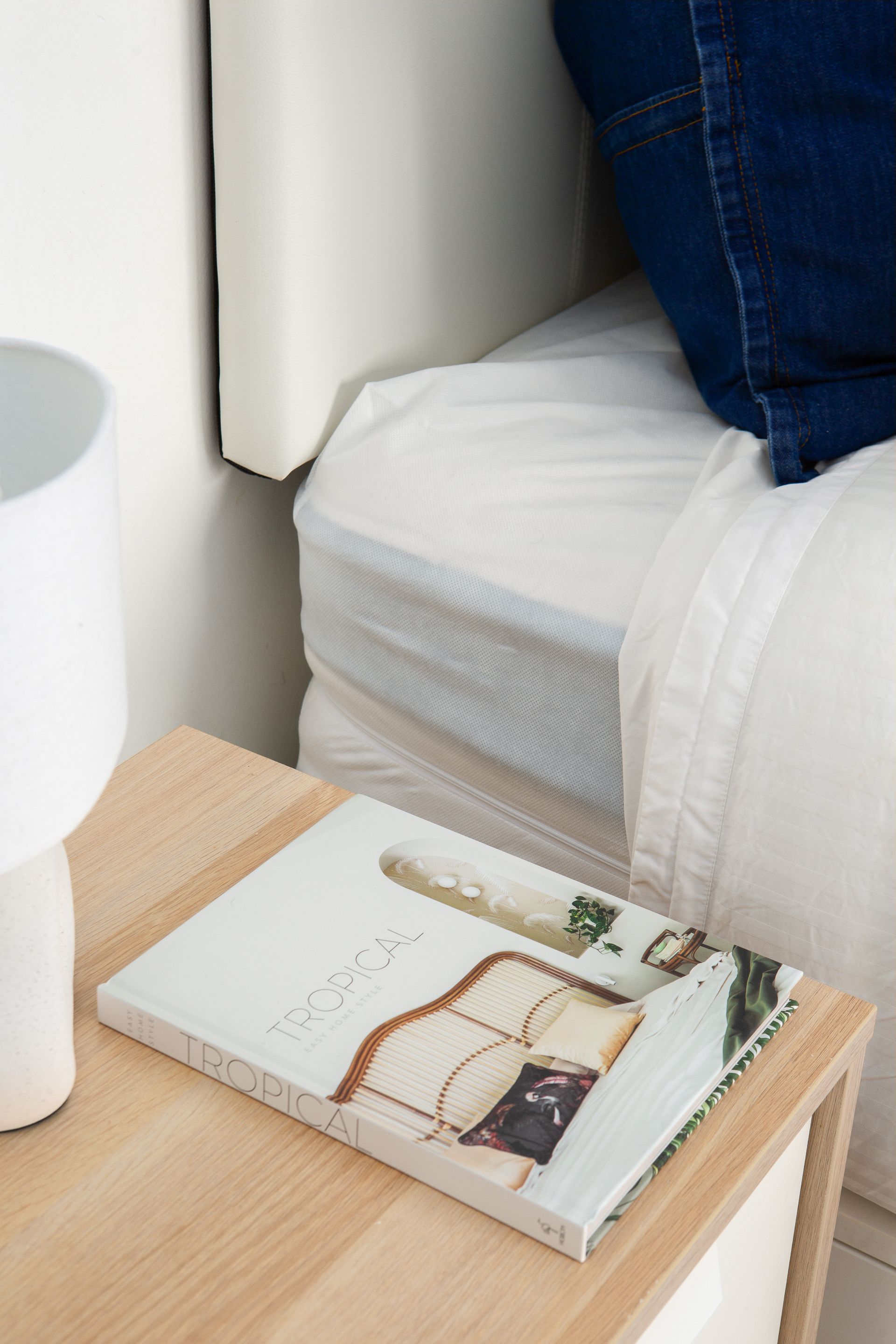 White bedside table with a magazine beside a bed and a denim-clad leg in a small bedroom corner