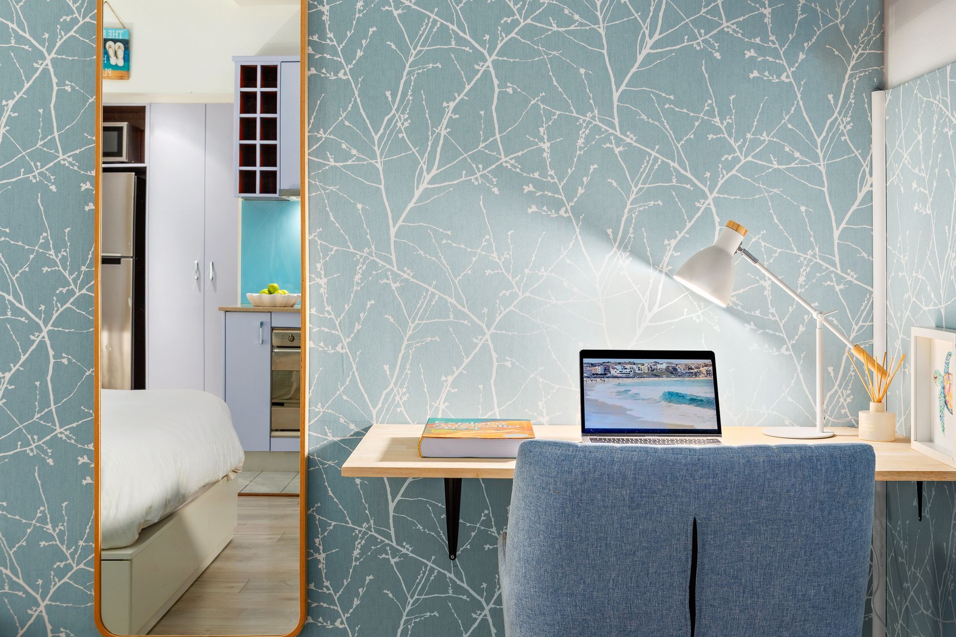 Bright home office with laptop, desk lamp, blue chair, and tree-patterned wallpaper beside a bedroom doorway