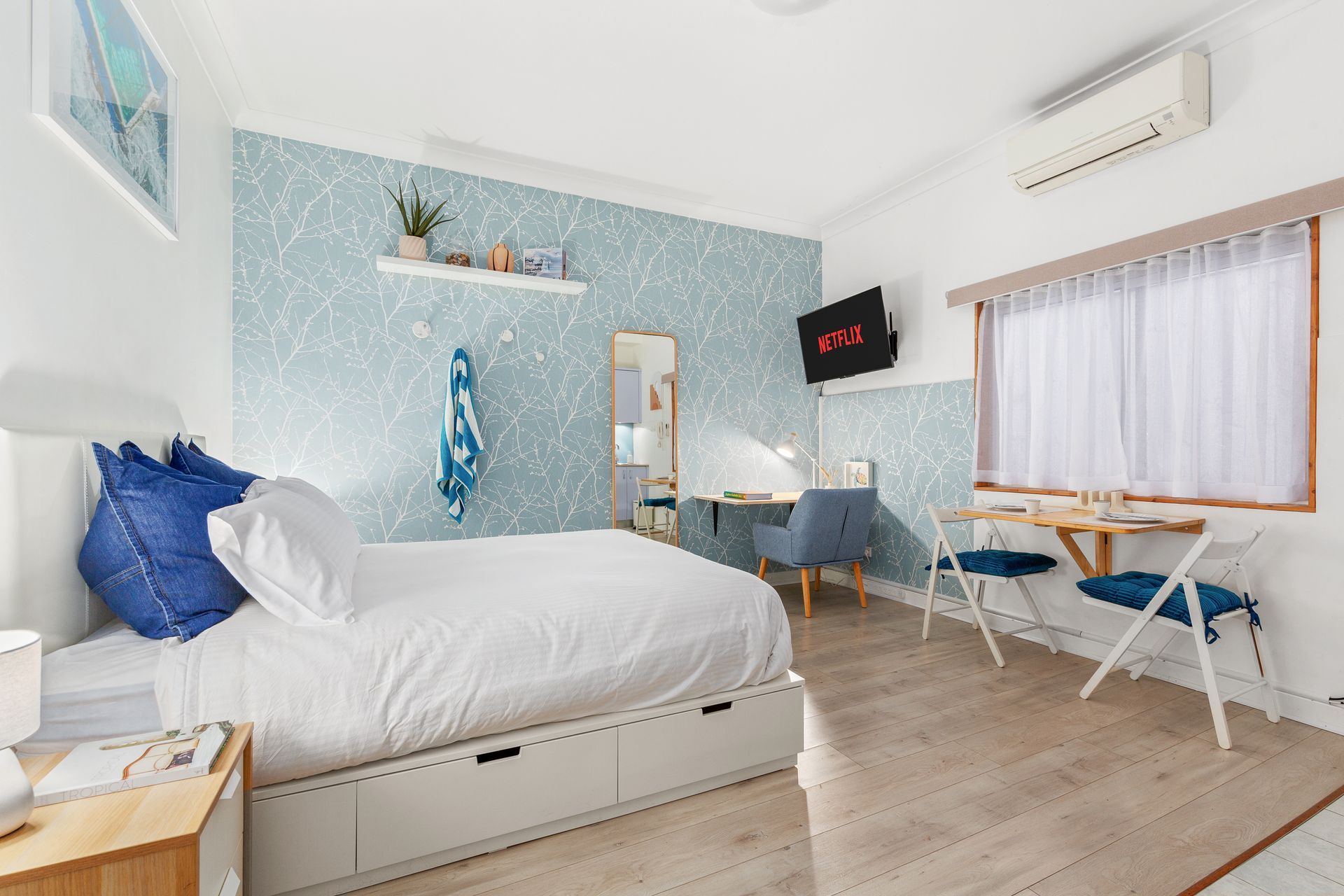 Bright modern bedroom with a white bed, blue accents, desk, and window seating.