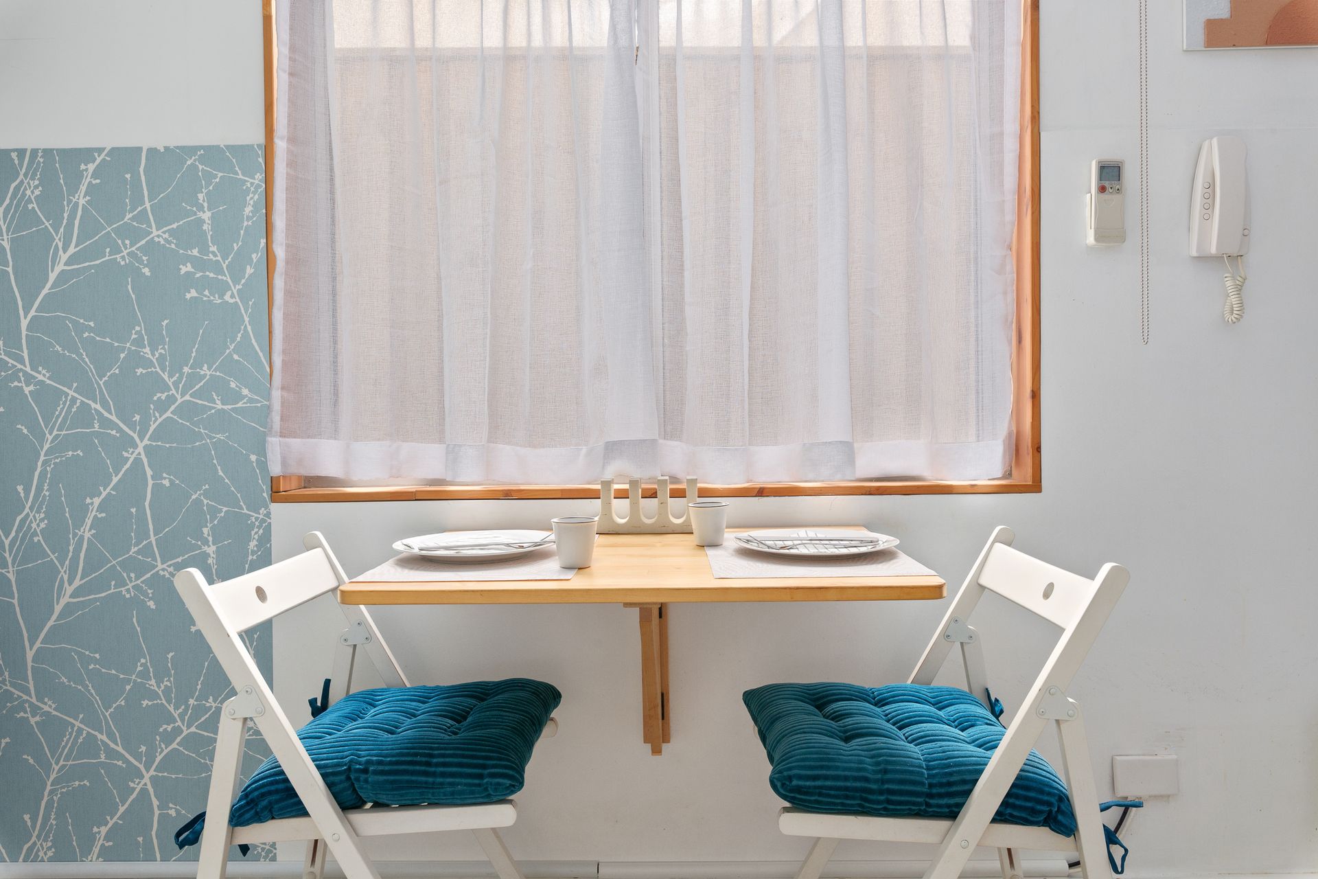 Small dining nook with a wooden table, white chairs, and blue cushions beside a window and sheer curtains.