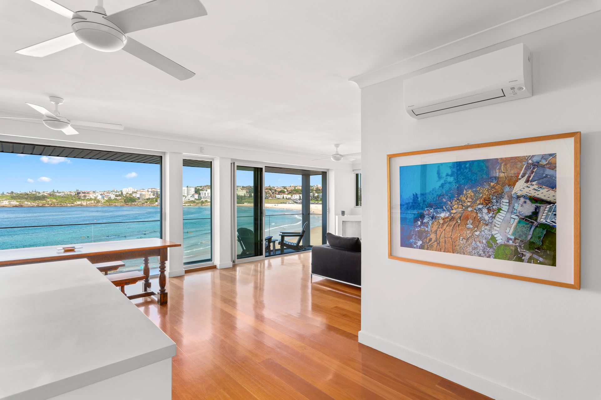 Bright ocean-view living room with hardwood floors, ceiling fan, and framed artwork on white walls