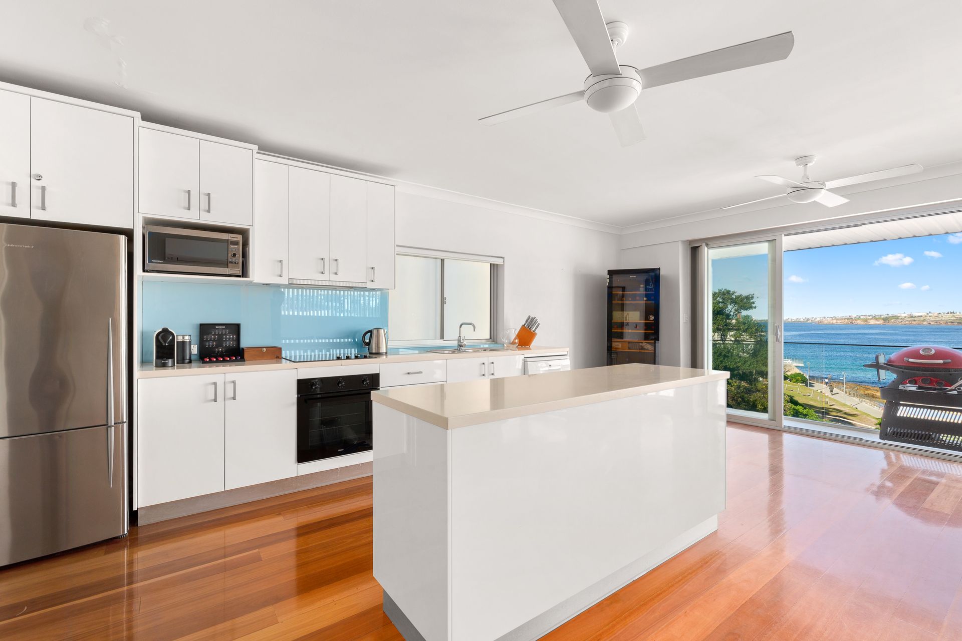 Bright modern kitchen with white cabinets and island, opening to a sunlit ocean-view living area.