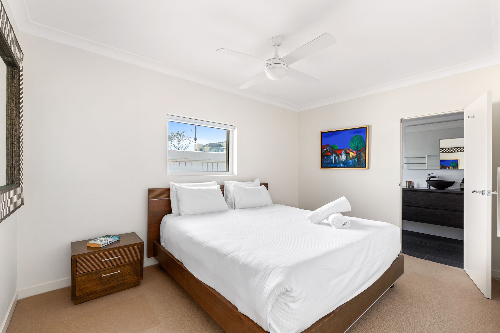 Bright bedroom with white bed, wicker chest, wall art, and open doorway to adjoining room.