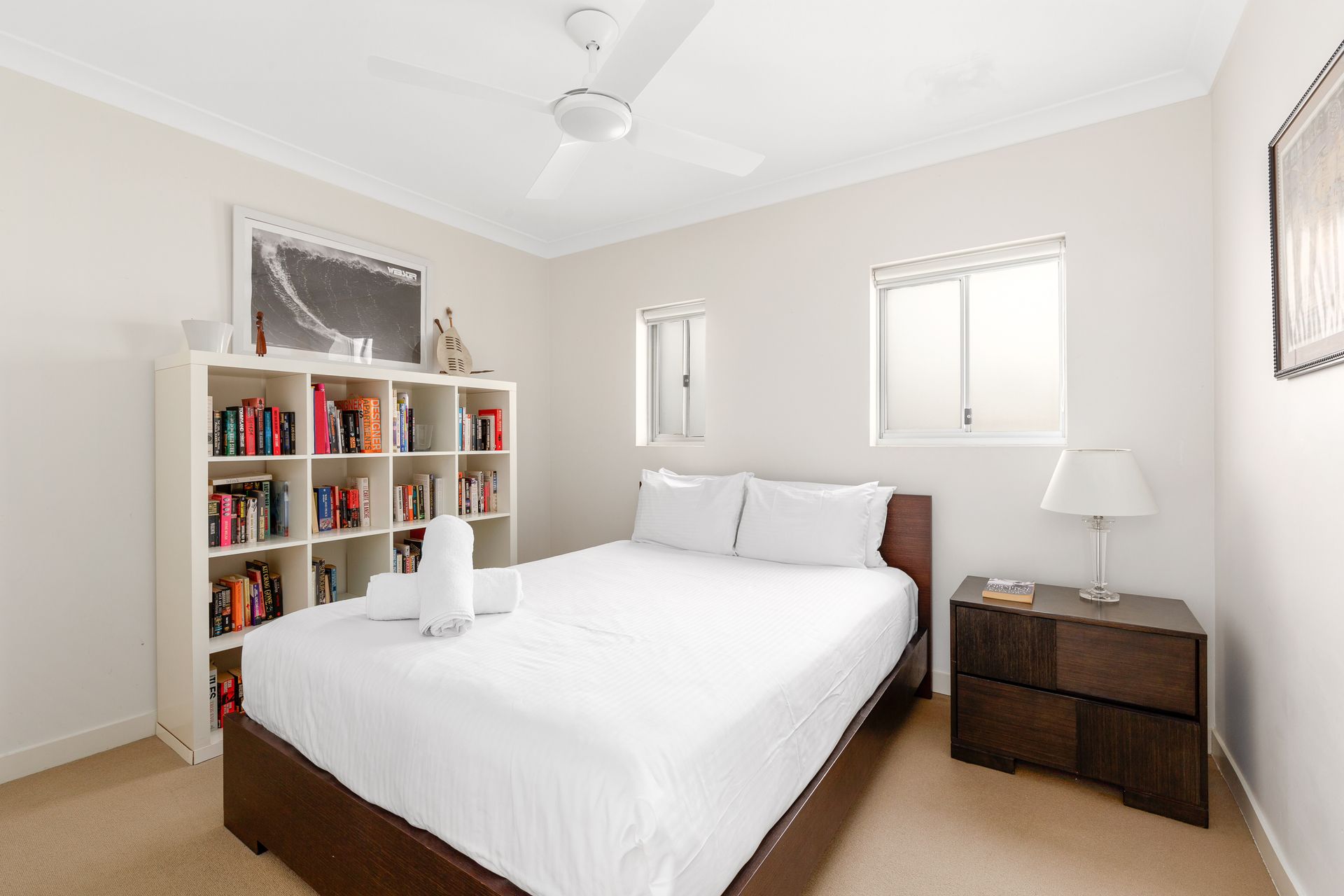 Bright bedroom with a white bed, bookshelf, nightstand, and two windows
