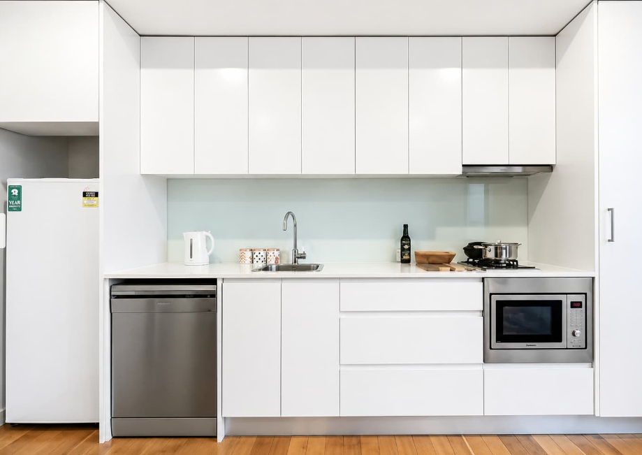 Modern white kitchen with cabinets, stainless dishwasher, microwave, refrigerator, and sink on a wooden floor