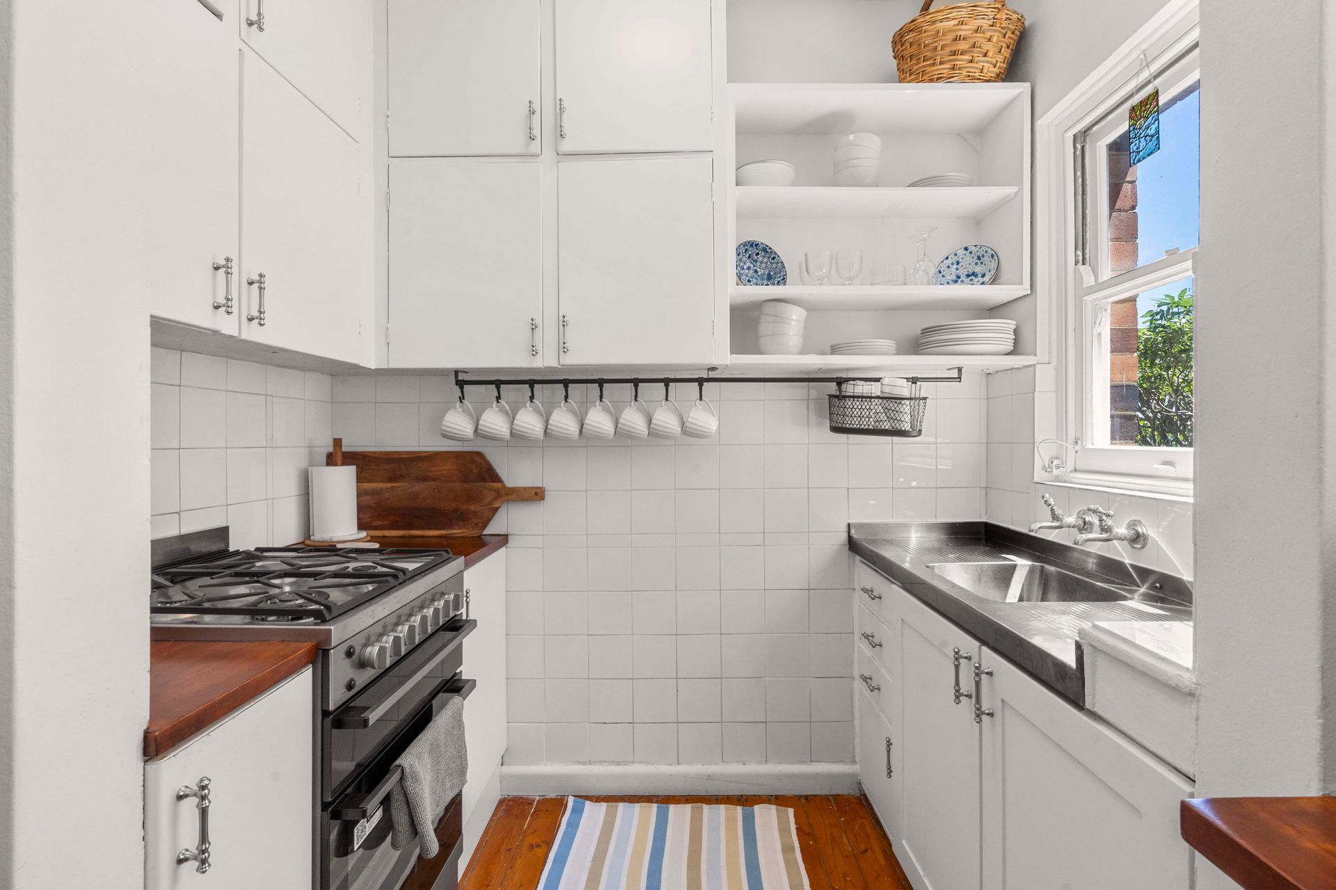Bright white galley kitchen with stove, sink, open shelves, and a colorful striped rug