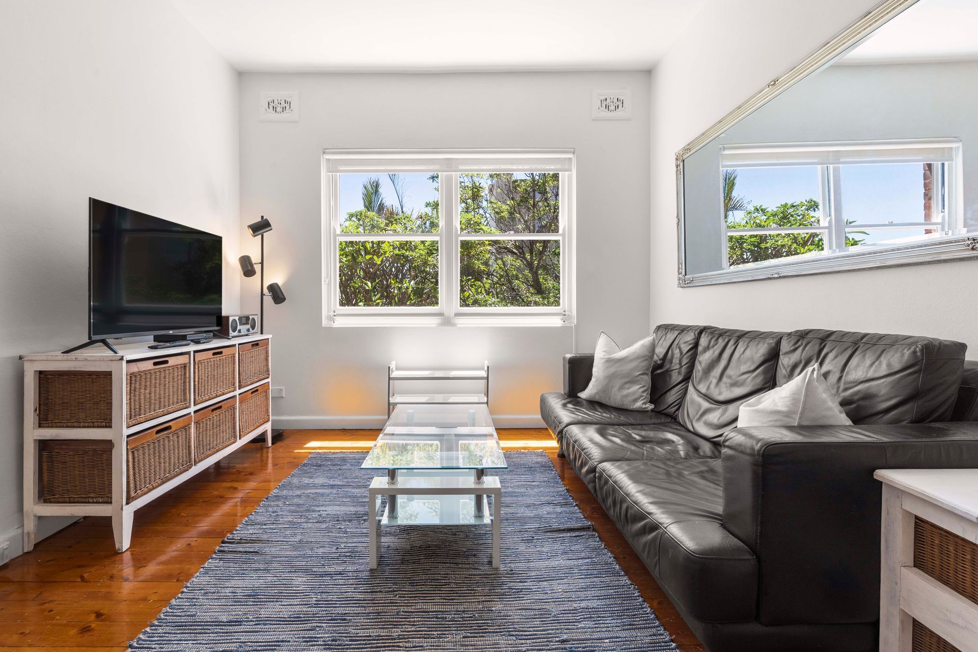 Modern living room with black leather sofa, TV, wicker cabinet, glass coffee table, and bright windows