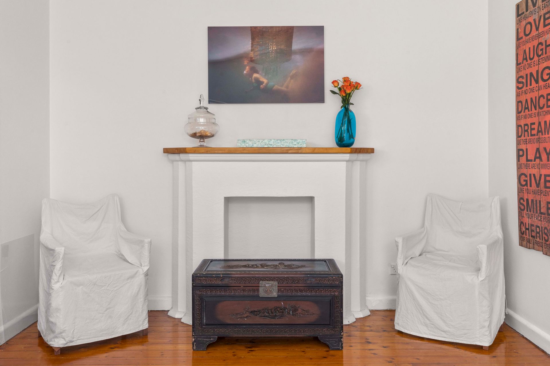 Living room with two white armchairs, a dark chest, fireplace, and flowers on the mantel.