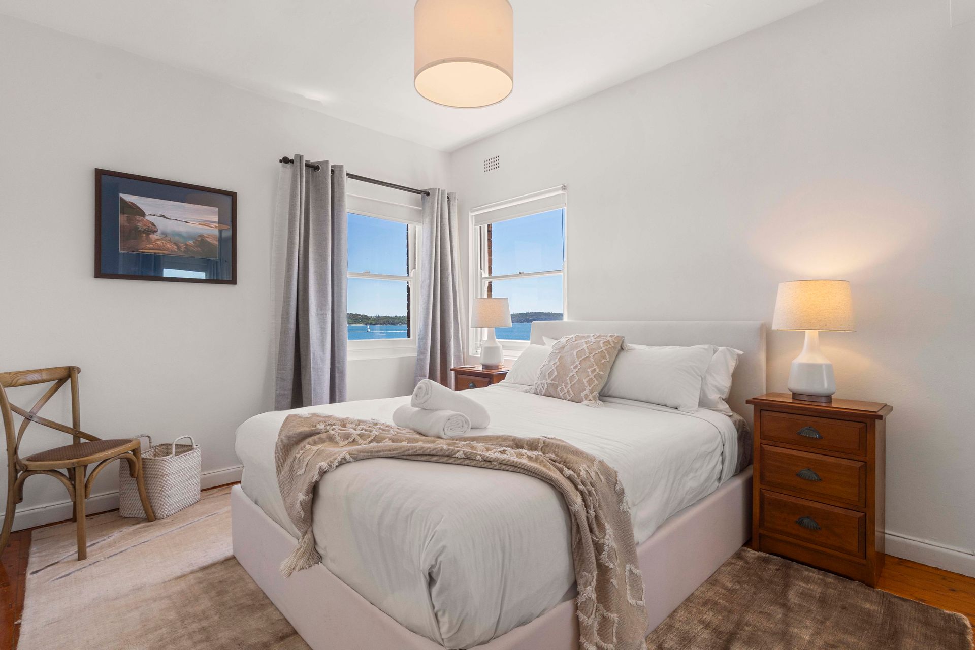 Bright bedroom with a made bed, bedside lamp, chair, and ocean view through windows