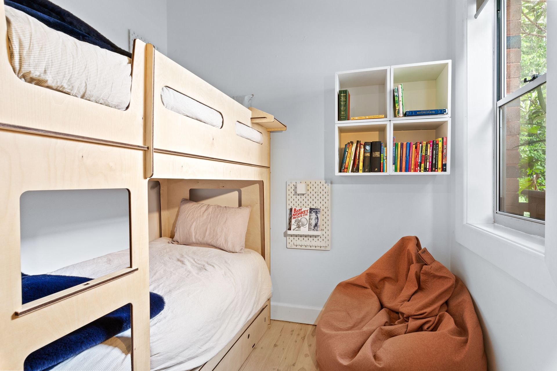 Small bunk bedroom with white walls, a bookshelf, bean bag chair, and a window view of greenery.