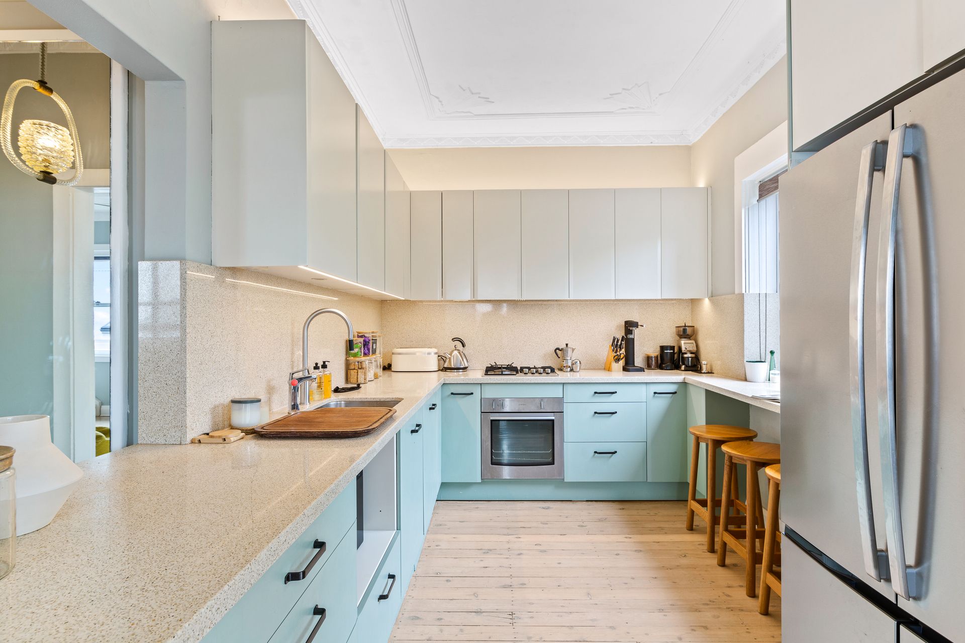 Bright kitchen with light blue cabinets, white countertops, stainless fridge, and a stool by the window.