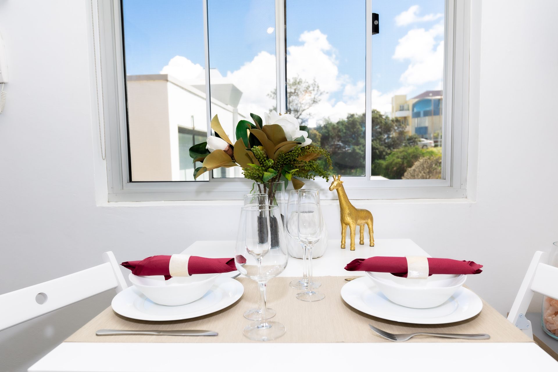 Set dining table by a bright window with white plates, red napkins, glassware, flowers, and giraffe figurines