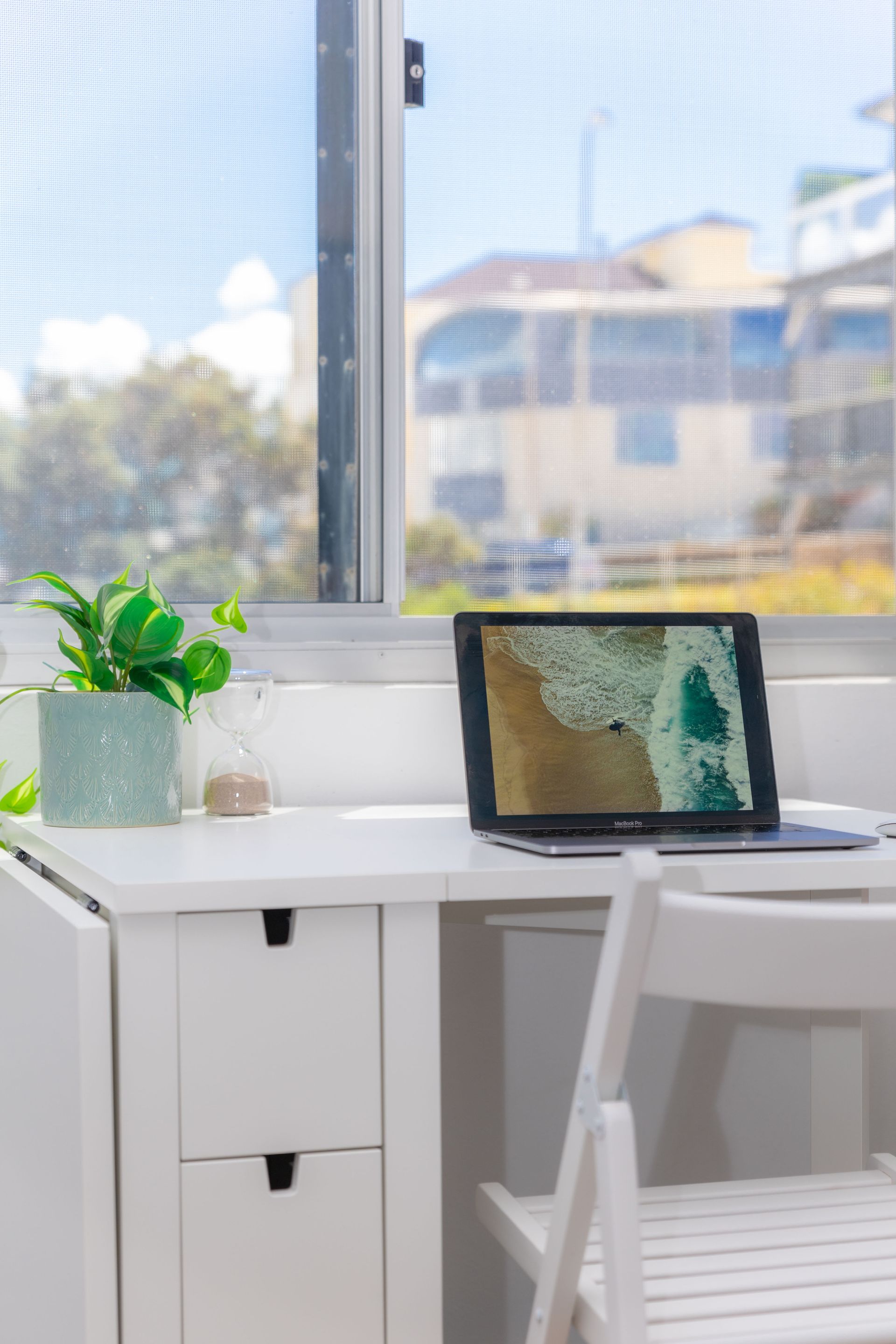 White desk by a sunny window with a laptop, potted plant, and chair in a bright room