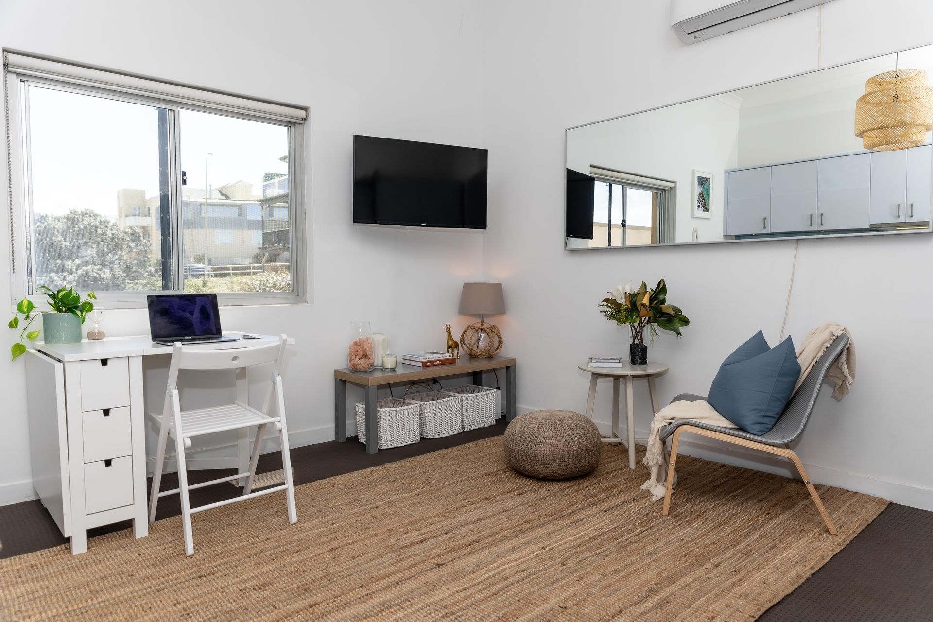 Bright living room with wicker chair, white desk by window, wall-mounted TV, and woven rug