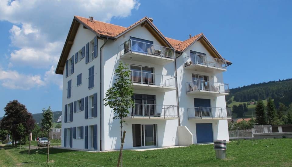 Immeuble blanc à plusieurs étages avec balcons, volets bleus et toit de tuiles rouges, se détachant sur un fond de verdure et de collines.