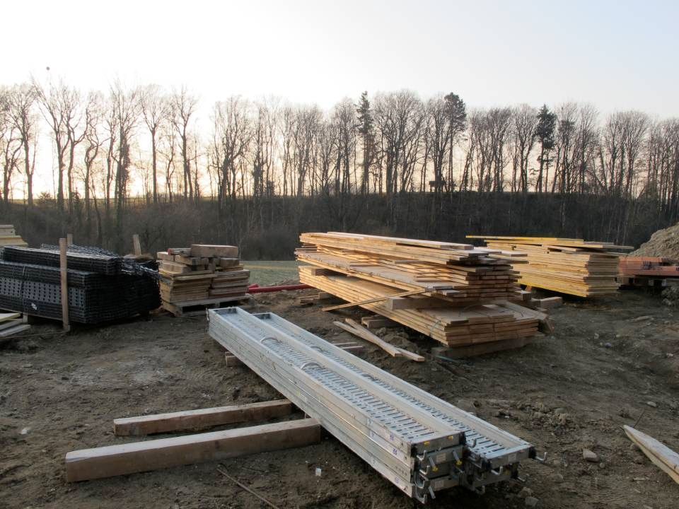Des piles de bois et de poutres métalliques sur un chantier de construction, des arbres en arrière-plan, au crépuscule.