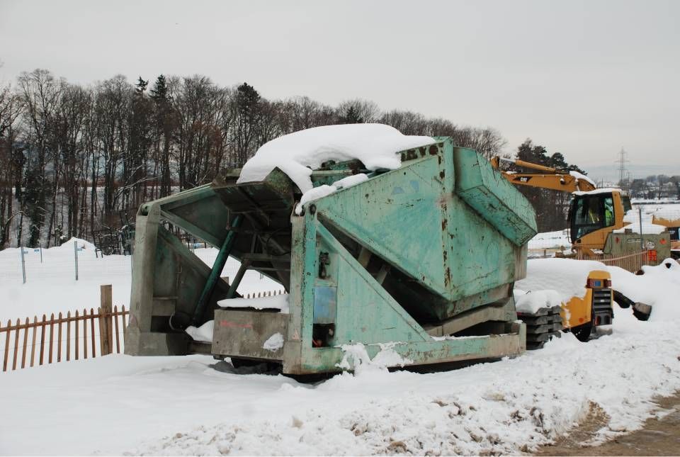 Grande structure industrielle de couleur turquoise recouverte de neige, probablement sur un chantier. Une pelleteuse jaune est visible en arrière-plan.