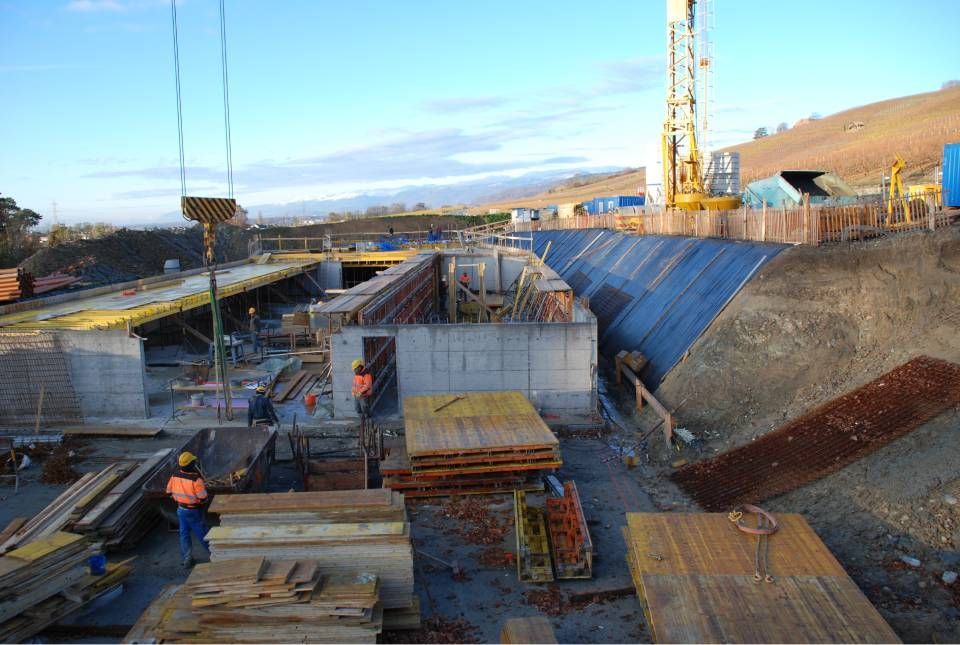 Chantier de construction avec structures en béton, grue et ouvriers ; flanc de colline en arrière-plan.