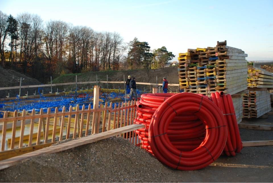 Chantier de construction avec tuyaux rouges, palettes en bois et ouvriers ; sol bleu et jaune.