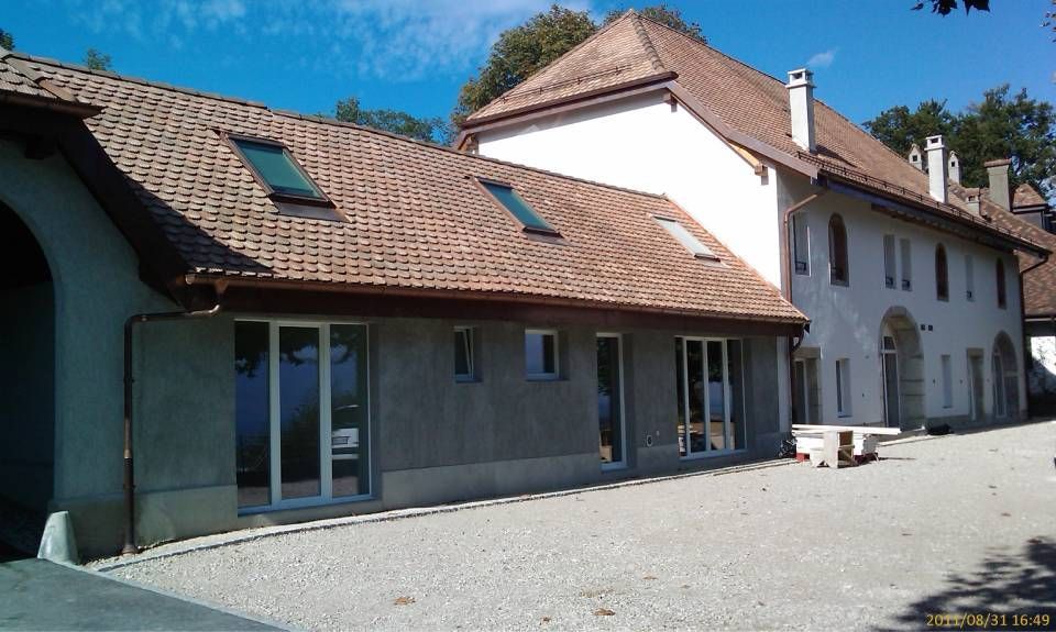 Bâtiment au toit de tuiles brunes, aux murs blancs et à la cour pavée, sous un ciel bleu.