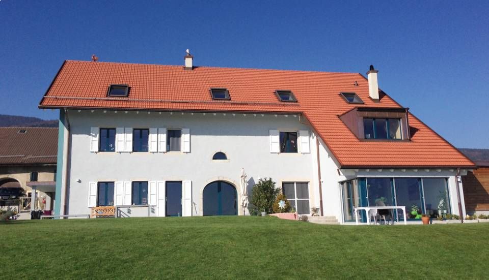 Maison blanche avec un toit de tuiles rouges, des puits de lumière et une véranda vitrée par une journée ensoleillée.
