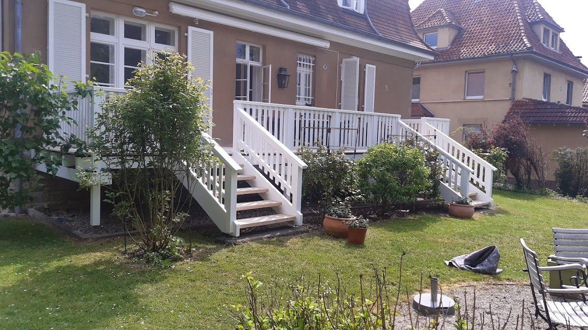 Weiße Terrasse und Treppe, die zu einem Haus mit braunen Wänden führen, umgeben von Grün.