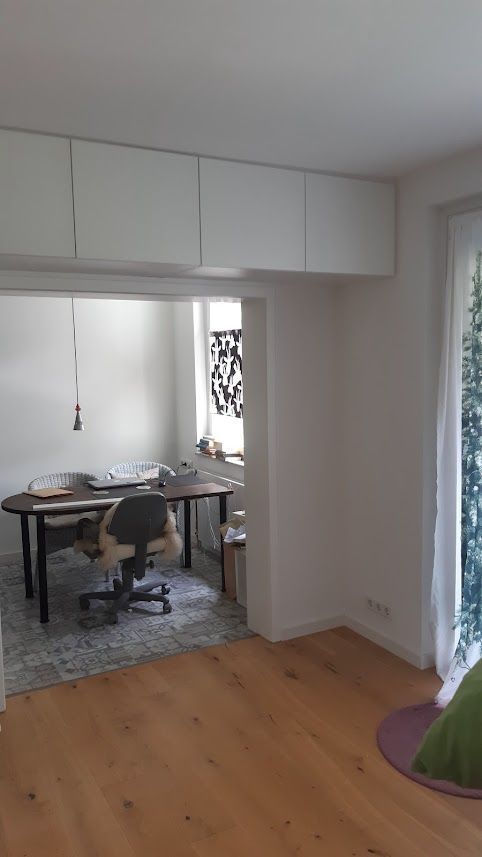 Büroraum: Weiße Hängeschränke, Schreibtisch mit Stuhl und Hartholzboden. Sonnenlicht durch das Fenster.