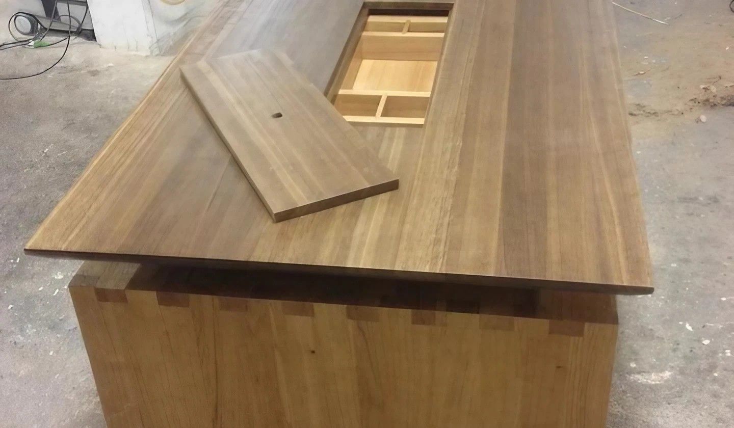Holztisch mit einem zentralen Fach, dessen Deckel teilweise geöffnet ist. Am Sockel ist eine Schwalbenschwanzverbindung sichtbar.