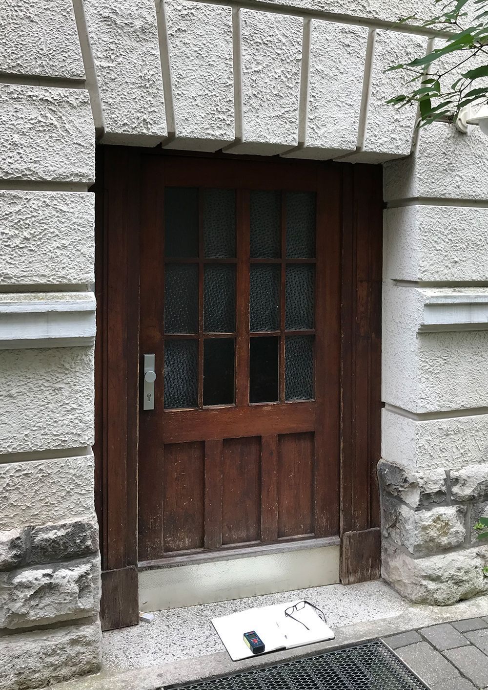 Holztür mit Glasscheiben in einer Steinfassade. Auf dem Boden liegen eine schwarze Fußmatte und ein kleiner Gegenstand.